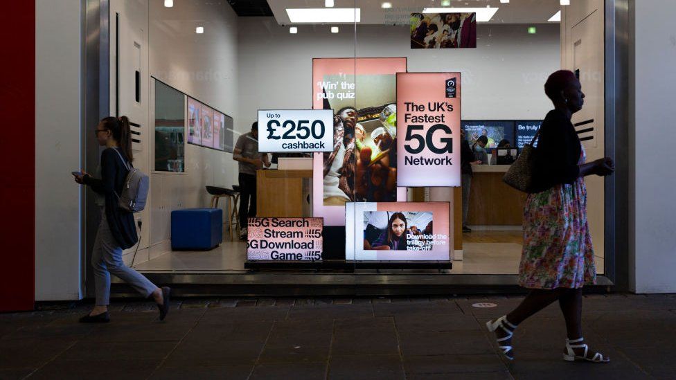 Pedestrians walk by a high street branch of Three UK displaying adverts for their 5G network